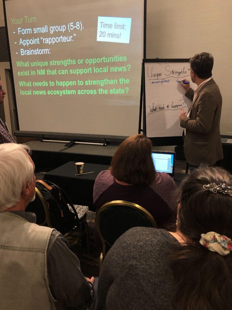A man wearing a blazer lists "unique strengths" and "resourcefulness" on an easel. Beside him is a slideshow of questions about local news in New Mexico.